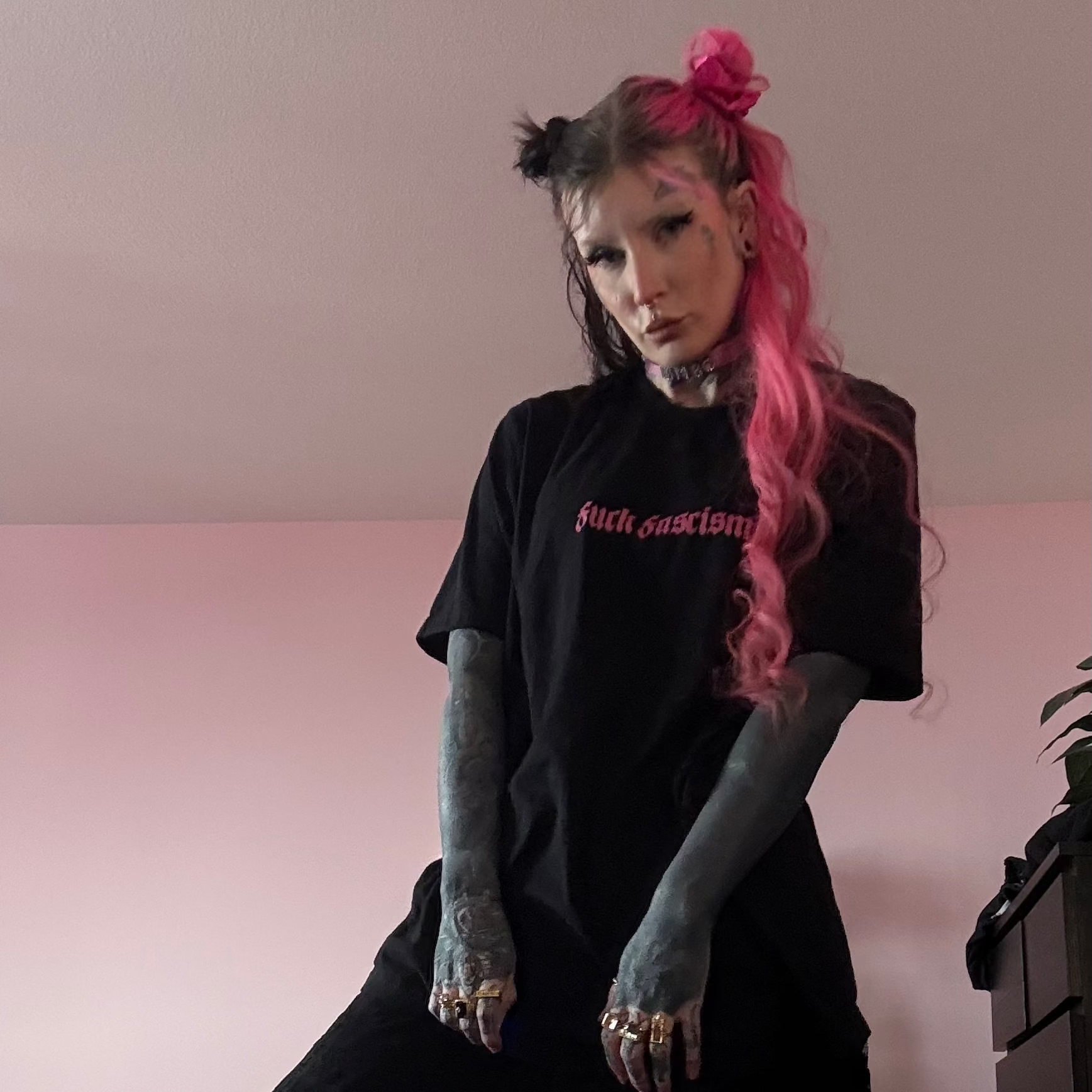 Person wearing a black outfit with text, standing against a pink wall.