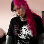 Person with pink hair wearing a black t-shirt with a graphic design, sitting indoors.