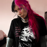 Person with pink hair wearing a black t-shirt with a graphic design, sitting indoors.