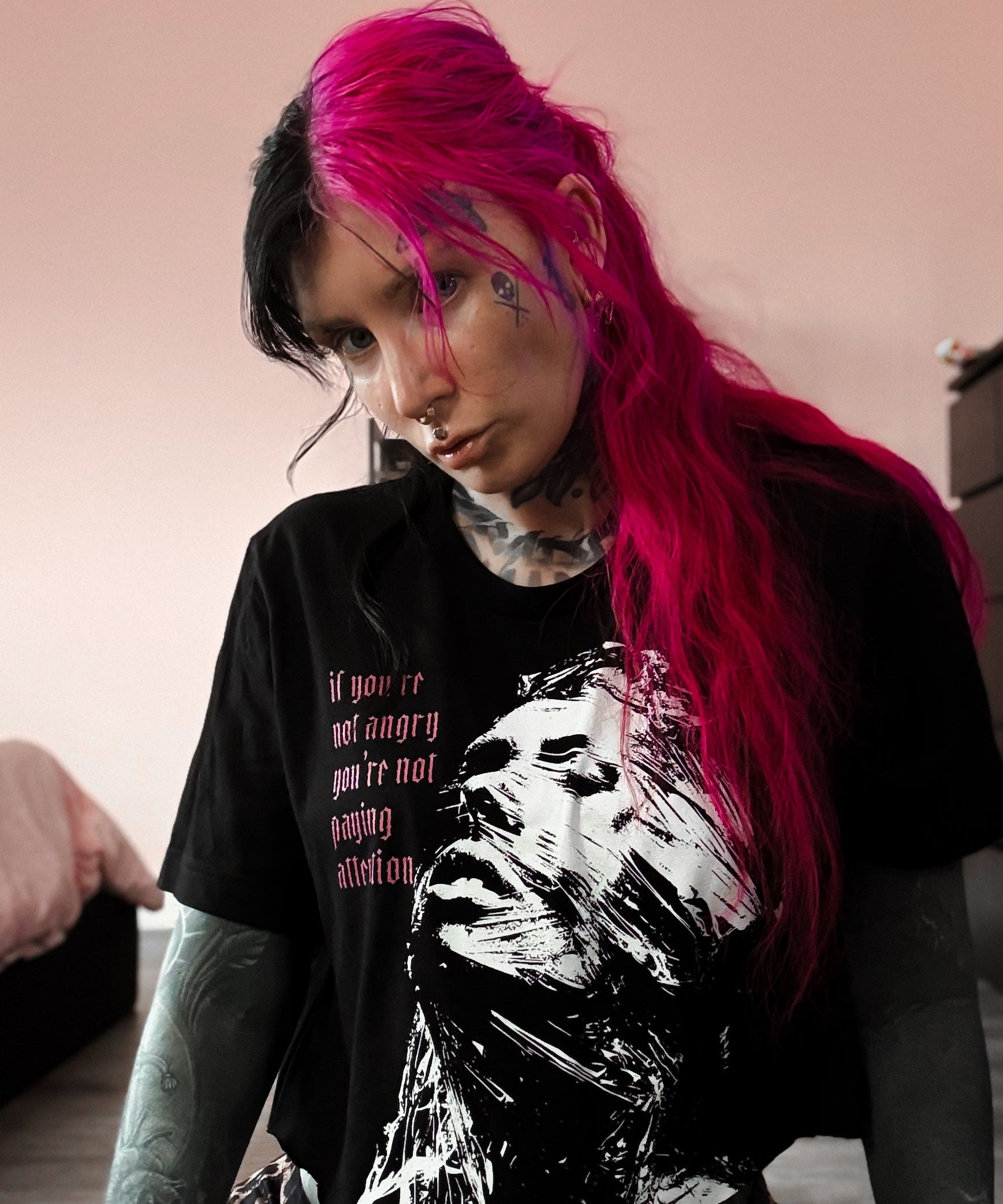Person with pink hair wearing a black t-shirt with a graphic design, sitting indoors.