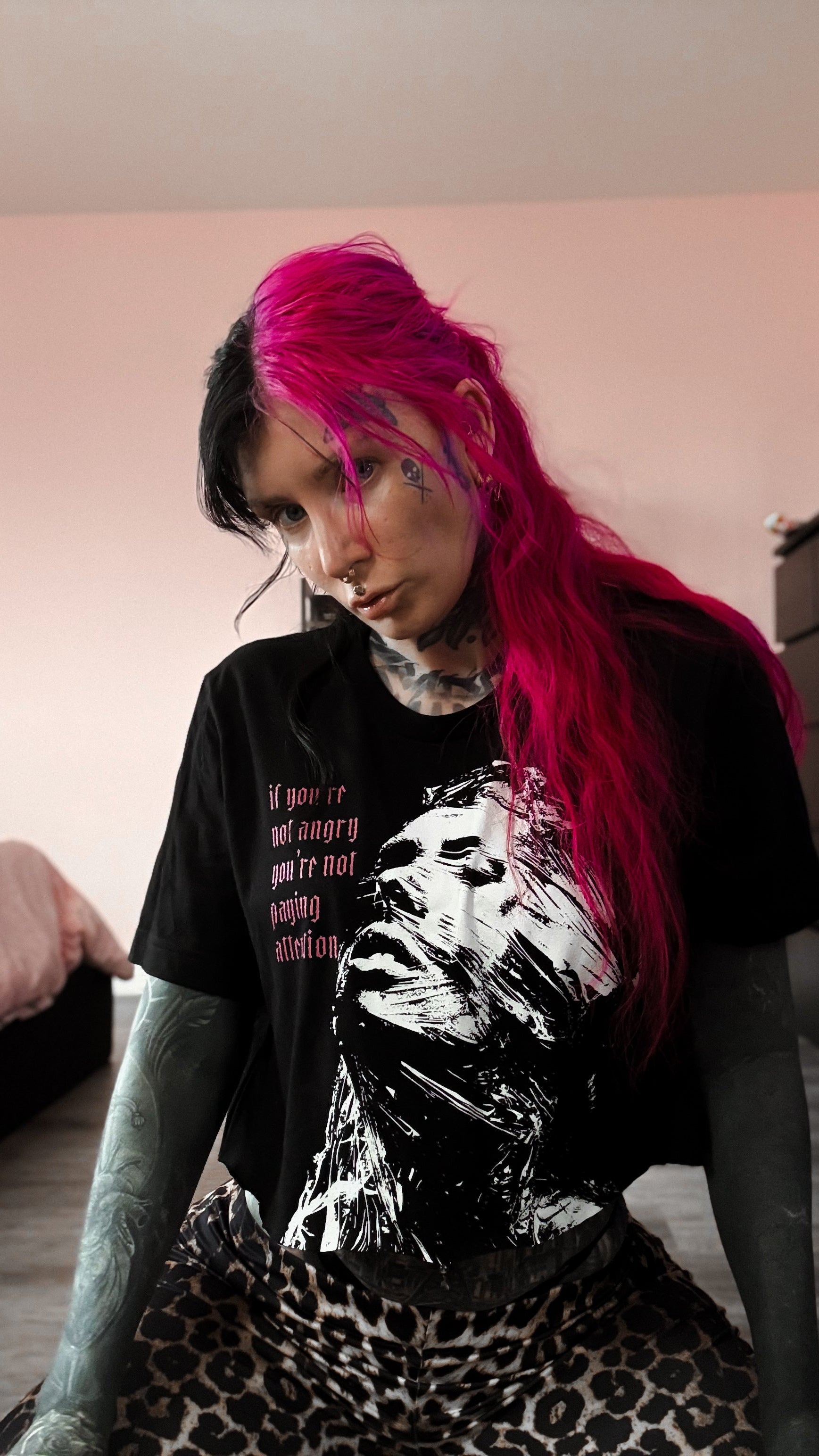 Person with pink hair wearing a black t-shirt with a graphic design, sitting indoors.