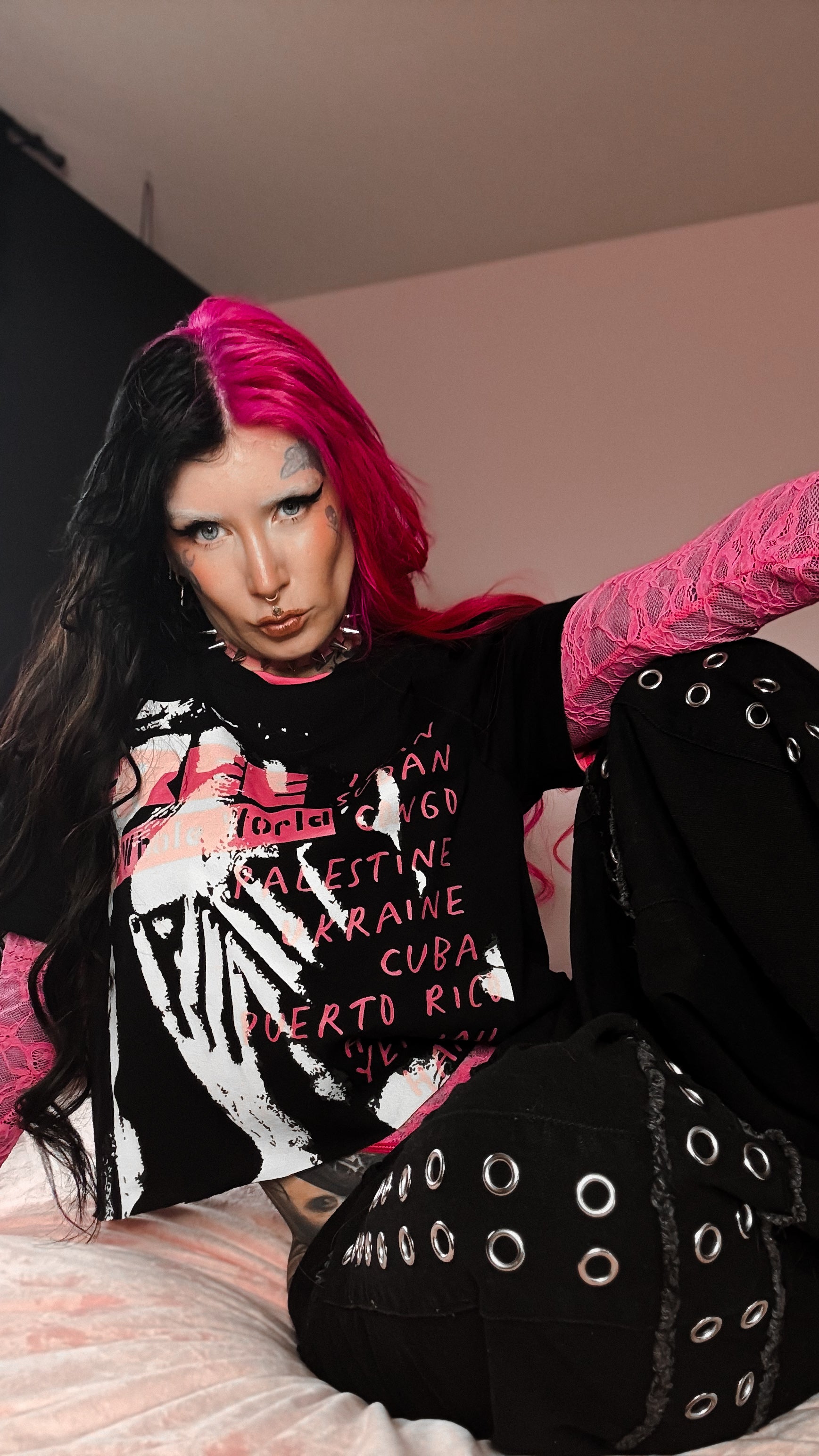 Person with pink hair and black outfit sitting on a bed
