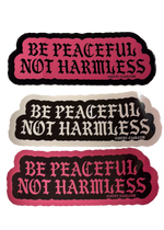 Three scalloped bumper stickers with the text 'BE PEACFUL NOT HARMLESS' in black gothic lettering. The top and bottom stickers have a pink background, and the middle sticker has a light beige background.