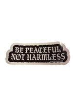 A die-cut sticker with the text 'BE PEACEFUL NOT HARMLESS' in white gothic-style lettering on a black background, 