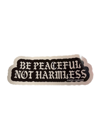 A die-cut sticker with the text 'BE PEACEFUL NOT HARMLESS' in white gothic-style lettering on a black background, 