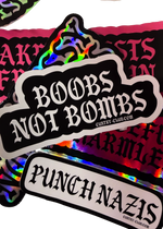 Black "Boobs Not Bombs" sticker with white gothic lettering on a pile of Cuntry Club stickers