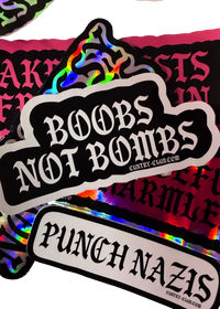 Black "Boobs Not Bombs" sticker with white gothic lettering on a pile of Cuntry Club stickers