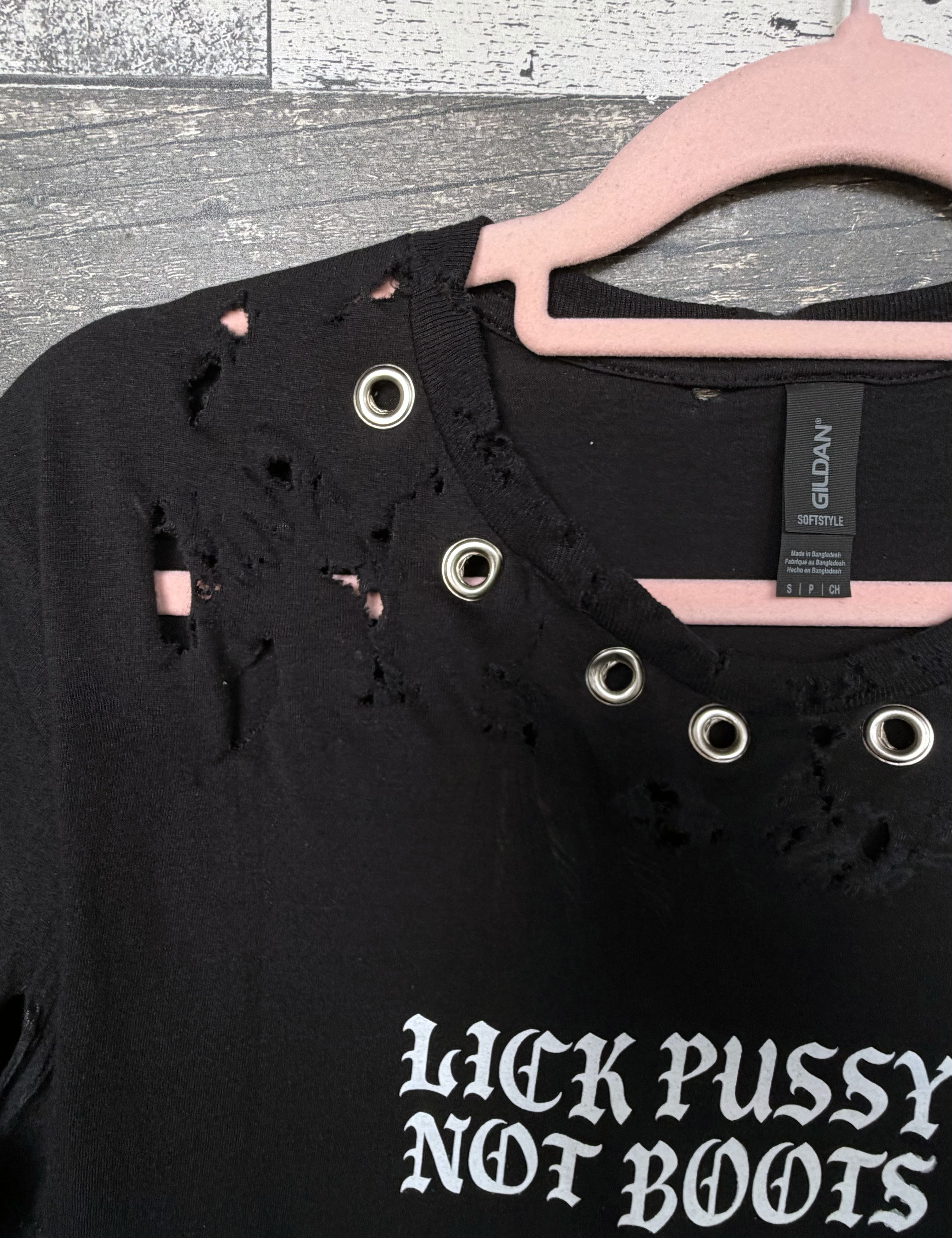 Black distressed shirt with 'LICK PUSSY NOT BOOTS' text on a hanger.